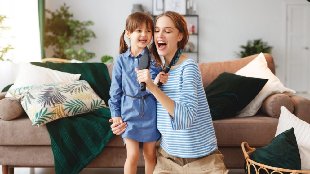 Mother and daughter, both with long brown hair, sing into brushes in their living room