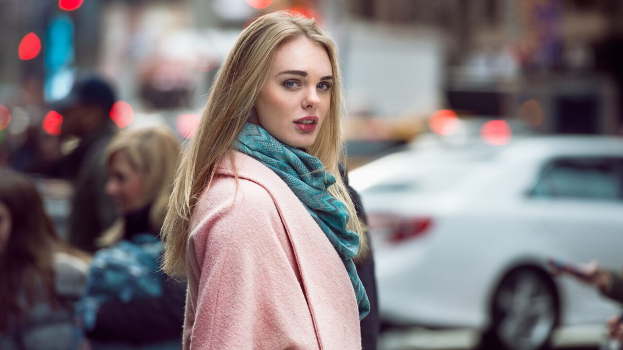 Beautiful elegant woman walking on the crowd on New York city street wearing pink fashionable jacket and scarf at winter time. Fashion model girl crossing the street.