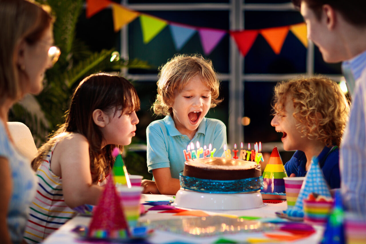 Child blowing candles on cake surrounded by rainbow decorations.